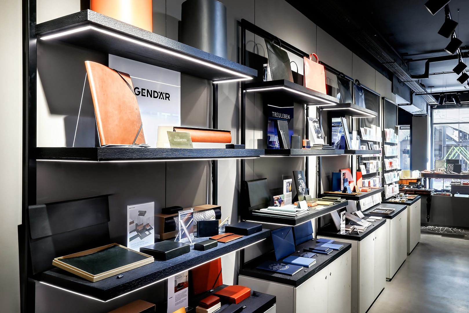 BEING BORN – Bethge Shop Hamburg: Legendär präsentiert auf beleuchteten Wandregalen Bethge Shop Hamburg – Retail Design von BEING BORN mit beleuchteten Wandregalen aus schwarzer Stahlkonstruktion und grauen, kubischen Elementen für Legendär.
